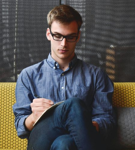 A young man in glasses writes in a notebook while sitting on a stylish couch indoors.